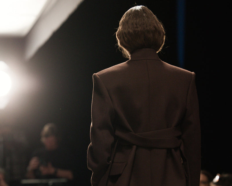 Model walking on a runway wearing a brown outfit with a blurred audience in the background